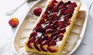 Pyszna tarta ze śliwkami, udekorowana wiórkami kokosowymi, prezentuje się apetycznie na srebrnej tacy. Obok leżą świeże śliwki.