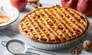 Złocista tarta z jabłkami, udekorowana kratką z ciasta, obok czerwonych jabłek i laski cynamonu.