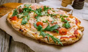 Pyszna pinsa z szynką parmeńską, rukolą i pomidorkami koktajlowymi, posypana parmezanem.