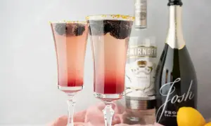 Dwa eleganckie drinki z prosecco, ozdobione jeżynami i złotym cukrem na krawędzi kieliszków. Obok butelka prosecco i wódki.