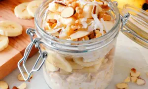 Słoik z nocną owsianką z bananem, posypaną płatkami kokosa i orzechami. Obok leżą plasterki banana i migdały.