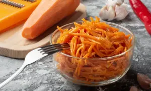 Świeża surówka z marchewki, przygotowana na tarce, czeka w szklanej misce na widelcu. Obok leżą czosnek i papryczka chili.