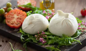 Dwie kulki burraty na rukoli, obok pomidorów i oliwy. To jest burrata – świeży ser z mozzarelli z kremowym wnętrzem.