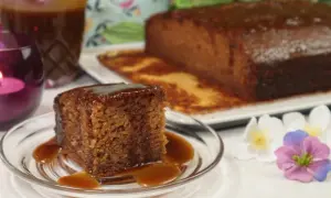 Pyszny kawałek sticky toffee pudding polany sosem karmelowym, obok cały bochenek i dzbanek sosu.