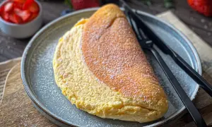 Puszysty omlet na talerzu, posypany cukrem pudrem. Idealny omlet przepis na pyszne śniadanie. Obok miseczka z truskawkami.