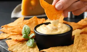 Chrupiące nachos zanurzane w kremowym, serowym dipie. Obok kawałek sera i listek bazylii. Idealna przekąska na imprezę.