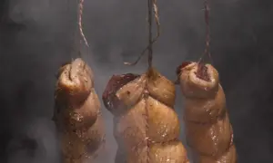 Trzy kawałki mięsa, zawieszone na sznurkach, w dymie. Szybkie peklowanie mięsa do wędzenia w toku.