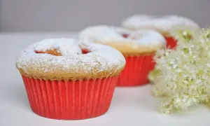 Pyszne muffinki dla dzieci, posypane cukrem pudrem, w czerwonych papilotkach, obok delikatnych białych kwiatów.
