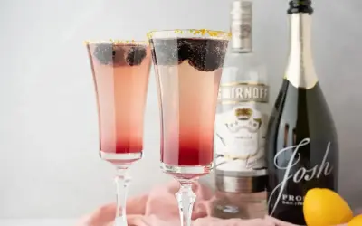 Dwa eleganckie drinki z prosecco, ozdobione jeżynami i złotym cukrem na krawędzi kieliszków. Obok butelka prosecco i wódki.