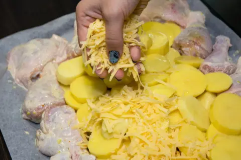 Ręka sypie startym serem na kurczaka i ziemniaki. Pyszny obiad z piekarnika gotowy do pieczenia.