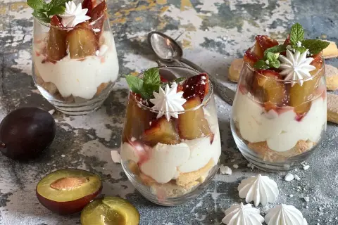 Pyszny deser ze śliwkami w pucharkach, przełożony kremem i biszkoptami, ozdobiony miętą i bezą.