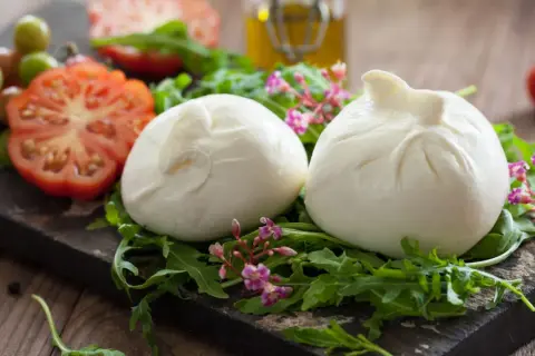 Dwie kulki burraty na rukoli, obok pomidorów i oliwy. To jest burrata – świeży ser z mozzarelli z kremowym wnętrzem.