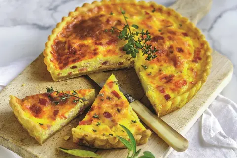 Złocisty quiche lorraine, pokrojony na kawałki, z boczkiem i ziołami, na drewnianej desce.