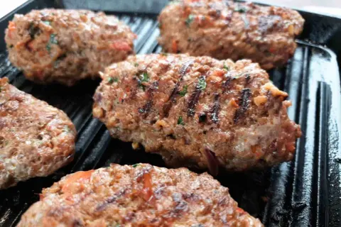Kebab Adana grillowany na patelni grillowej, z widocznymi kawałkami pomidorów i ziół.