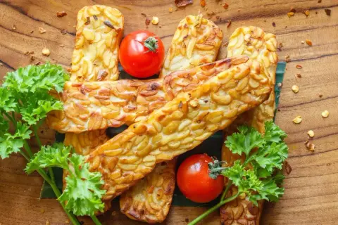 Usmażony tempeh, czyli co to jest? Złociste kawałki fasoli, pomidorki koktajlowe i natka pietruszki na drewnianej desce.