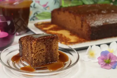 Pyszny kawałek sticky toffee pudding polany sosem karmelowym, obok cały bochenek i dzbanek sosu.