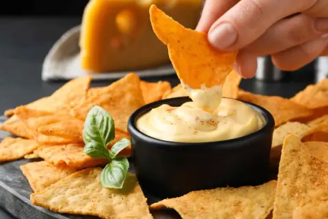 Chrupiące nachos zanurzane w kremowym, serowym dipie. Obok kawałek sera i listek bazylii. Idealna przekąska na imprezę.