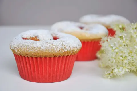 Pyszne muffinki dla dzieci, posypane cukrem pudrem, w czerwonych papilotkach, obok delikatnych białych kwiatów.