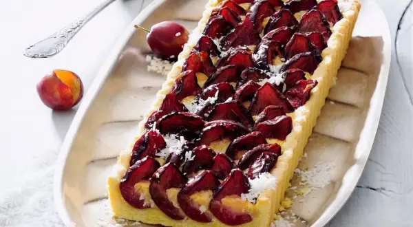 Pyszna tarta ze śliwkami, udekorowana wiórkami kokosowymi, prezentuje się apetycznie na srebrnej tacy. Obok leżą świeże śliwki.