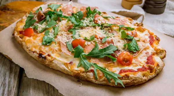 Pyszna pinsa z szynką parmeńską, rukolą i pomidorkami koktajlowymi, posypana parmezanem.