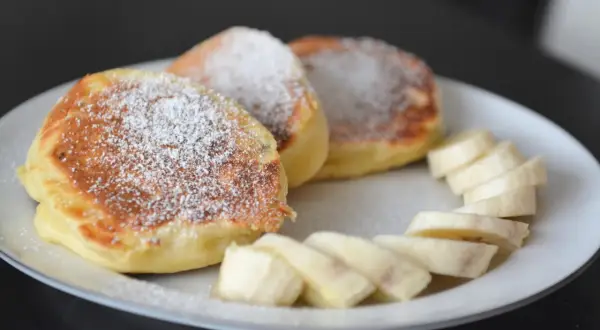 Puszyste, szybkie placuszki na śniadanie posypane cukrem pudrem, obok plasterki banana. Idealne na dobry początek dnia.