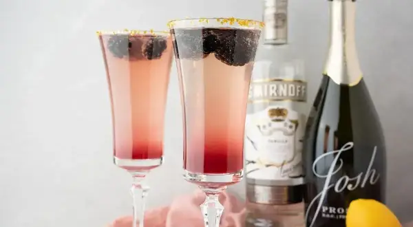 Dwa eleganckie drinki z prosecco, ozdobione jeżynami i złotym cukrem na krawędzi kieliszków. Obok butelka prosecco i wódki.