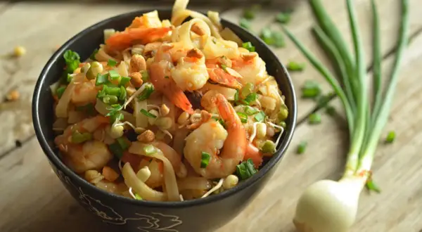 Aksamitne pad thai z krewetkami, posypane szczypiorkiem i orzeszkami, czeka w czarnej misce na drewnianym blacie. Obok leży cebulka.