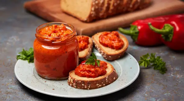 Słoik ajvaru i pieczywo z pastą. Ajvar do czego? Idealny na kanapki, jako dodatek do mięs czy warzyw.