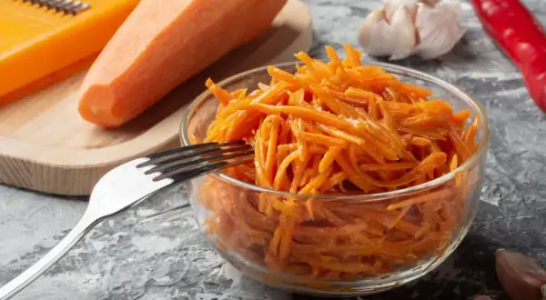 Świeża surówka z marchewki, przygotowana na tarce, czeka w szklanej misce na widelcu. Obok leżą czosnek i papryczka chili.
