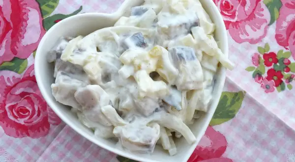 Pyszny przepis na śledzie w śmietanie z jabłkiem i ogórkiem. Sałatka w białej miseczce w kształcie serca na tle różowych róż.