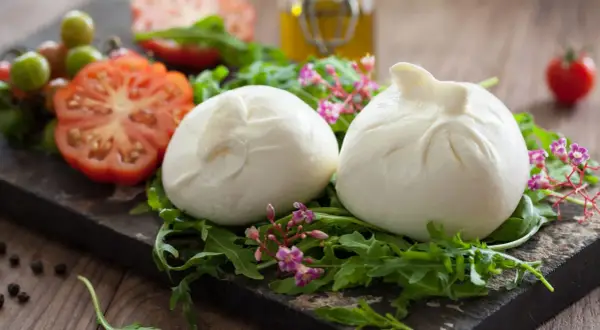 Dwie kulki burraty na rukoli, obok pomidorów i oliwy. To jest burrata – świeży ser z mozzarelli z kremowym wnętrzem.