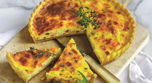 Złocisty quiche lorraine, pokrojony na kawałki, z boczkiem i ziołami, na drewnianej desce.