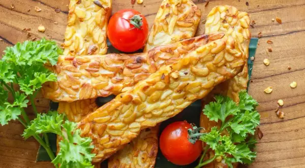 Usmażony tempeh, czyli co to jest? Złociste kawałki fasoli, pomidorki koktajlowe i natka pietruszki na drewnianej desce.