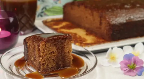 Pyszny kawałek sticky toffee pudding polany sosem karmelowym, obok cały bochenek i dzbanek sosu.