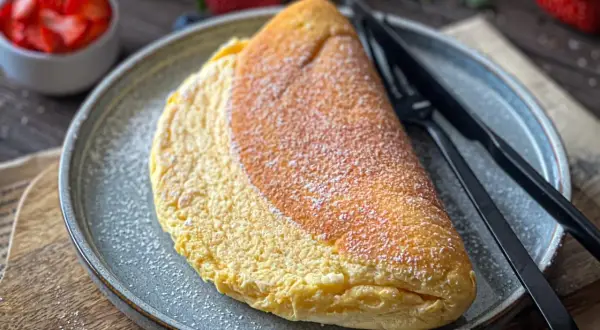 Puszysty omlet na talerzu, posypany cukrem pudrem. Idealny omlet przepis na pyszne śniadanie. Obok miseczka z truskawkami.