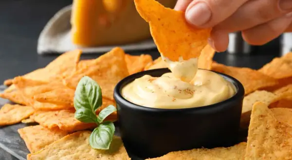 Chrupiące nachos zanurzane w kremowym, serowym dipie. Obok kawałek sera i listek bazylii. Idealna przekąska na imprezę.