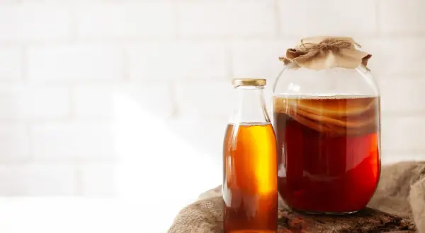 Słoik z ciemnym napojem i widocznym SCOBY, obok butelka z bursztynowym płynem. To jest co to jest kombucha, fermentowany napój.