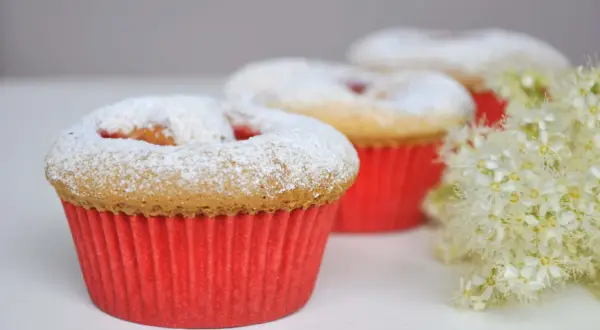 Pyszne muffinki dla dzieci, posypane cukrem pudrem, w czerwonych papilotkach, obok delikatnych białych kwiatów.