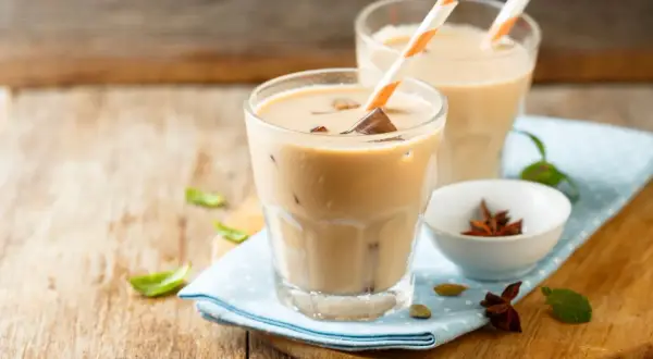 Dwa orzeźwiające **chai latte** z lodem i słomkami, na drewnianym blacie, obok miseczki z anyżem i kardamonem.
