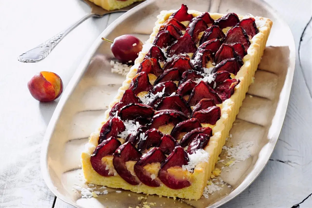 Pyszna tarta ze śliwkami, udekorowana wiórkami kokosowymi, prezentuje się apetycznie na srebrnej tacy. Obok leżą świeże śliwki.