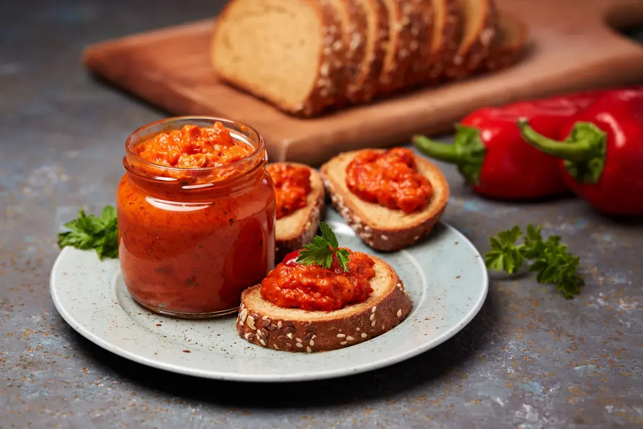 Słoik ajvaru i pieczywo z pastą. Ajvar do czego? Idealny na kanapki, jako dodatek do mięs czy warzyw.