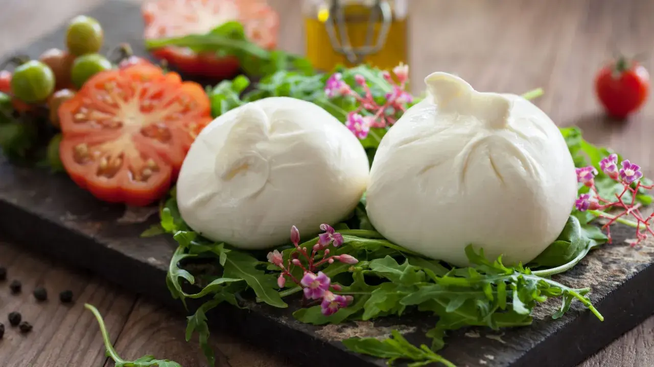 Dwie kulki burraty na rukoli, obok pomidorów i oliwy. To jest burrata – świeży ser z mozzarelli z kremowym wnętrzem.