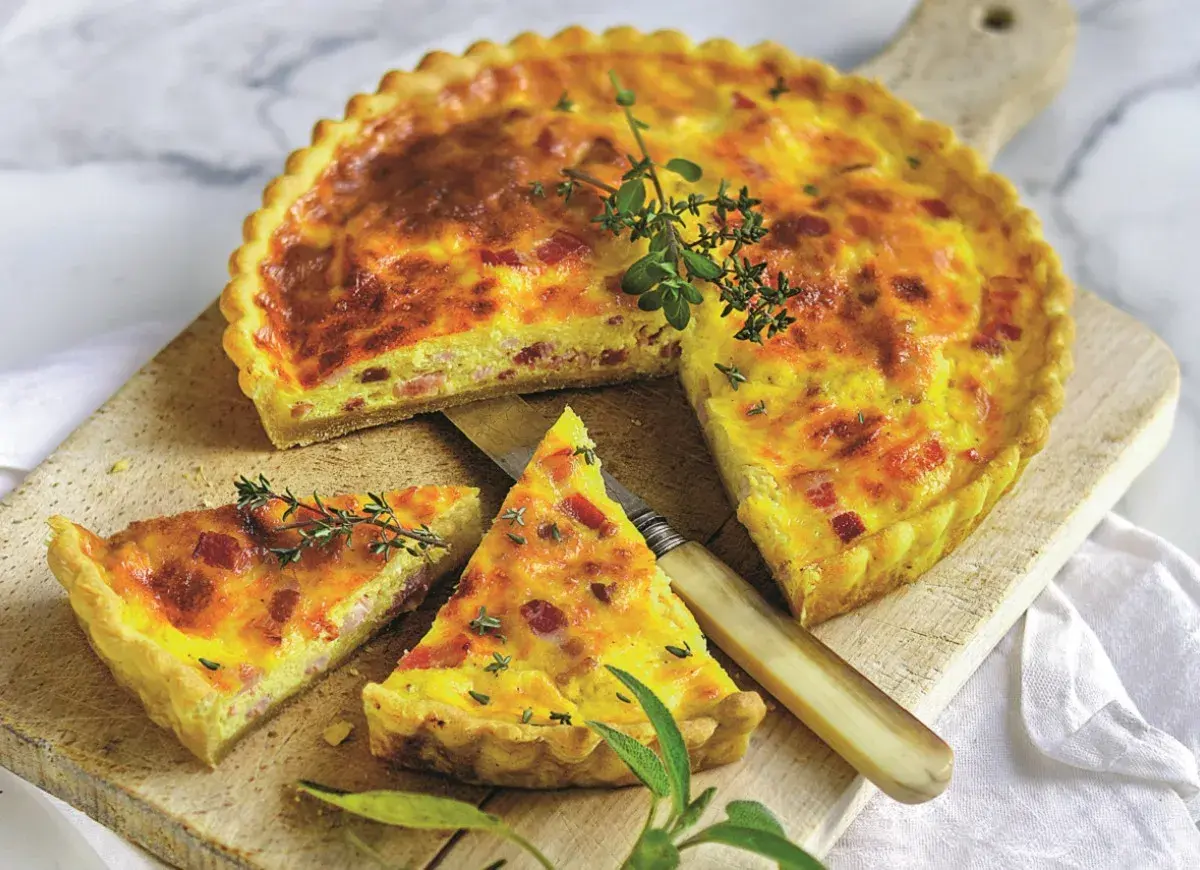Złocisty quiche lorraine, pokrojony na kawałki, z boczkiem i ziołami, na drewnianej desce.