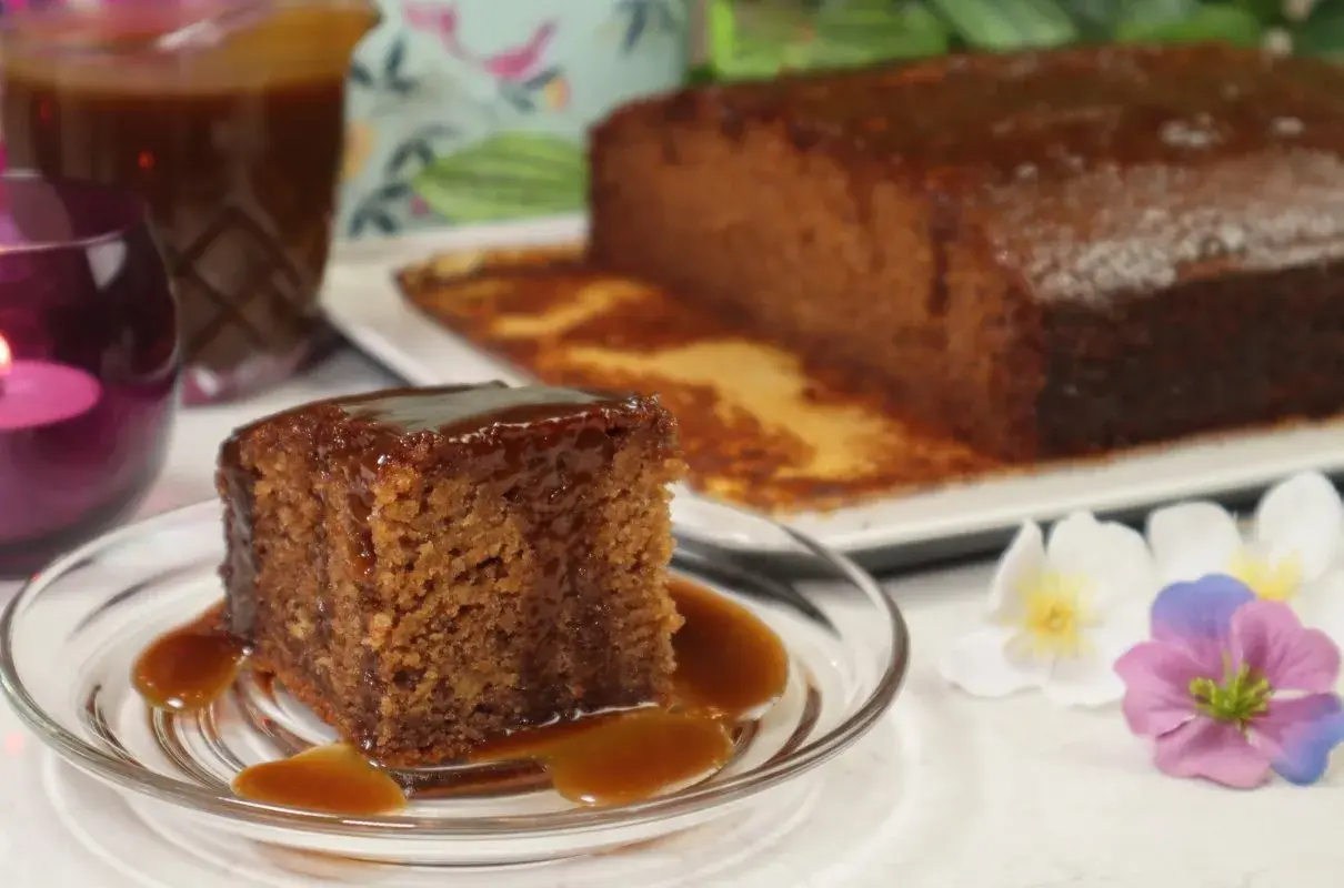 Pyszny kawałek sticky toffee pudding polany sosem karmelowym, obok cały bochenek i dzbanek sosu.