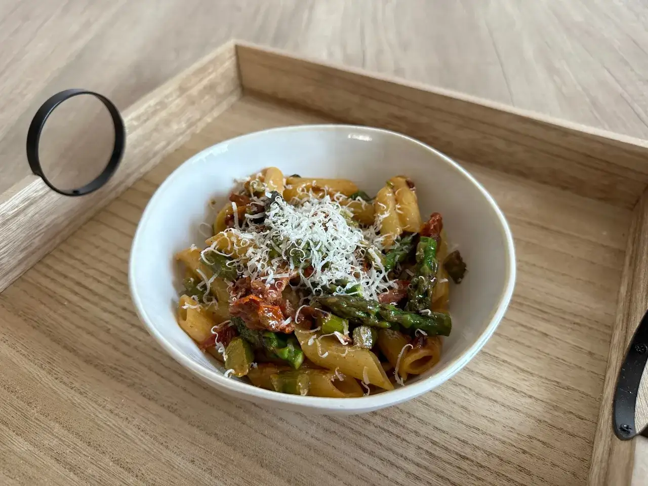 Penne z zielonymi szparagami i suszonymi pomidorami, posypane startym parmezanem, na drewnianej tacy.