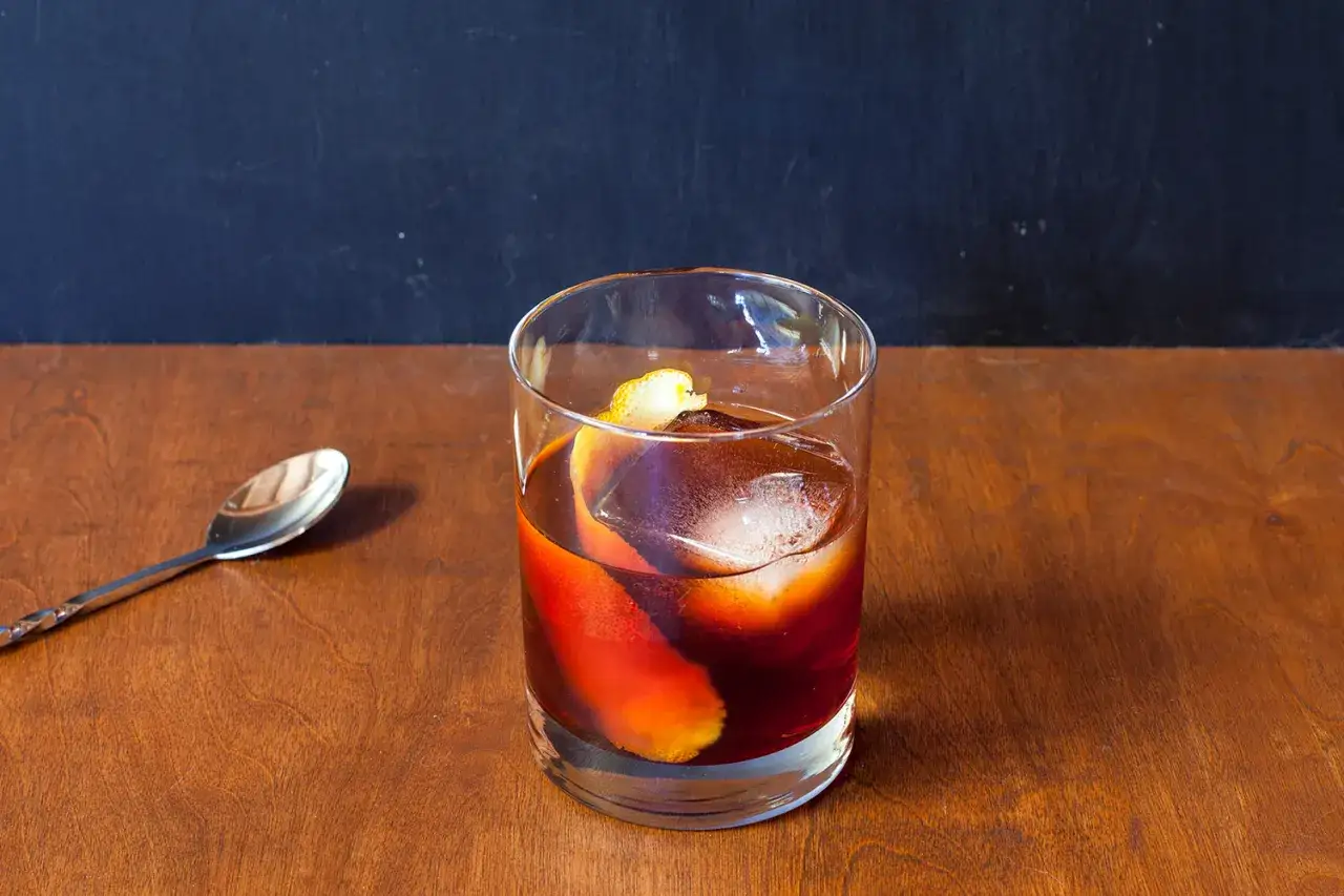 Klasyczny negroni przepis: czerwony koktajl z lodem i skórką pomarańczy w szklance, obok łyżeczka barmańska.