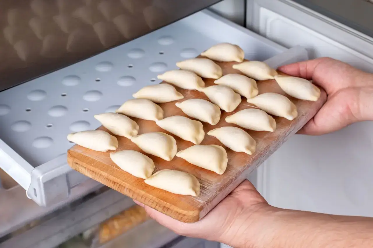 Ręce wkładają deskę z pierogami do zamrażarki. To prosty sposób, jak zamrozić ugotowane pierogi na później.