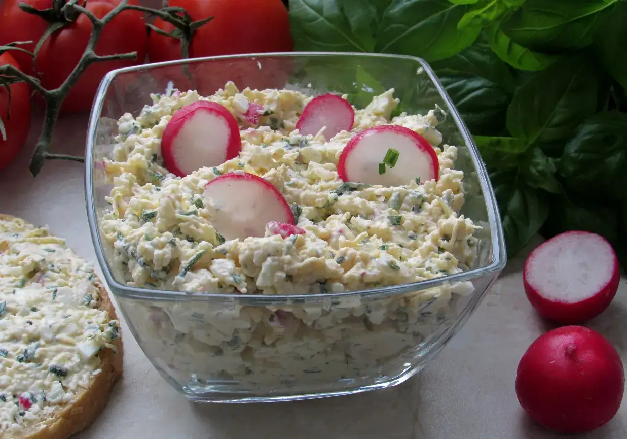 Pyszna pasta jajeczna z rzodkiewką i szczypiorkiem w szklanej misce, obok kromka chleba z pastą.