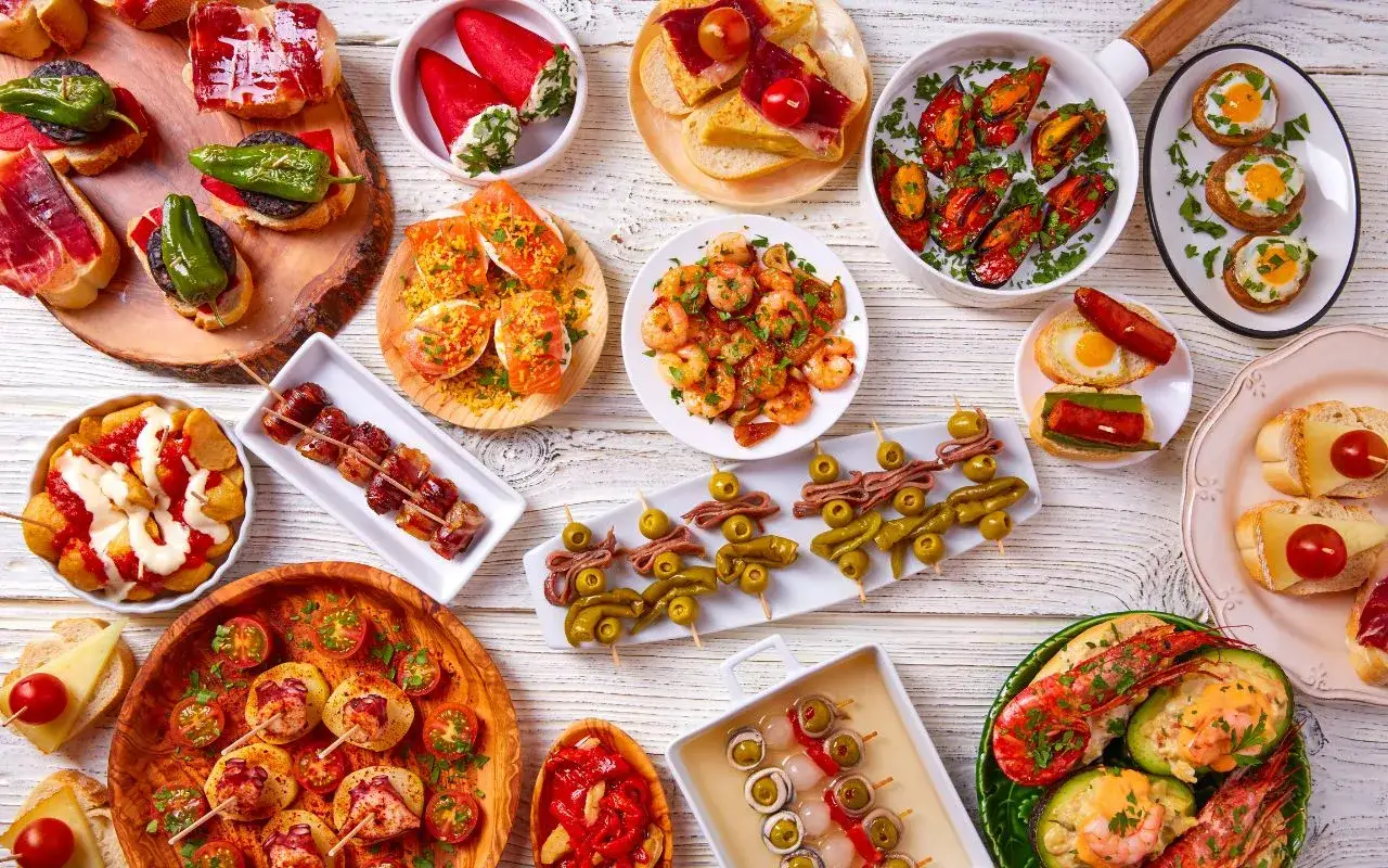 Tapas co to? Kolorowy stół pełen małych przekąsek: krewetki, małże, kanapki z szynką, oliwki, papryczki, jajka sadzone i inne smakołyki.