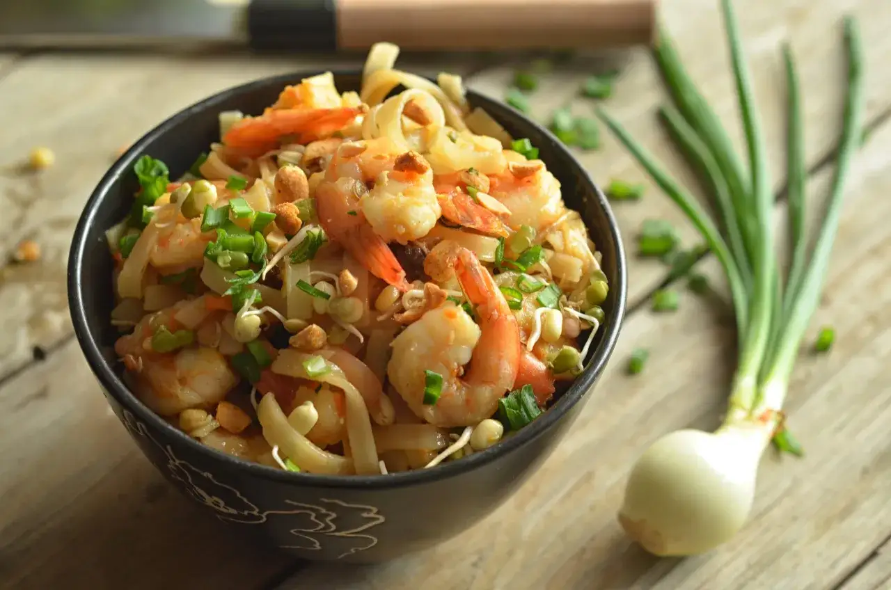 Aksamitne pad thai z krewetkami, posypane szczypiorkiem i orzeszkami, czeka w czarnej misce na drewnianym blacie.